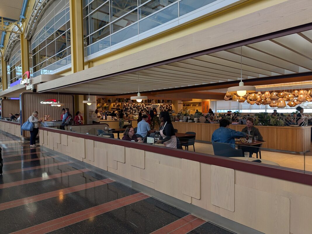 Capital One Venture X vs Amex Platinum: Is Half the Fee Enough? Capital One Landing at Reagan National Airport, an open mezzanine dining space with pale wood slat ceiling, pendant lights, travellers seated at tables and a food and drink counter behind