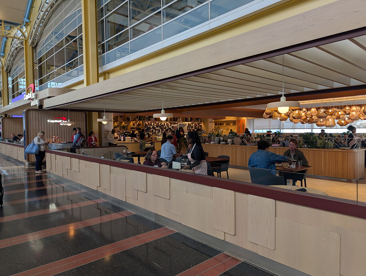 Capital One Landing at Reagan National Airport, an open mezzanine dining space with pale wood slat ceiling, pendant lights, travellers seated at tables and a food and drink counter behind