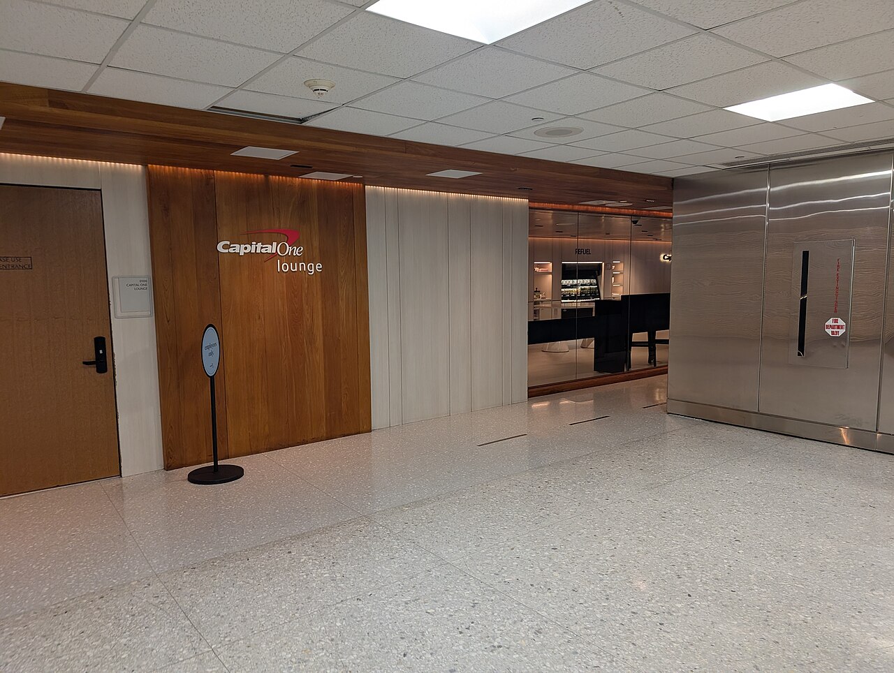 Entrance to the Capital One Lounge at Dulles International Airport, with the Capital One lounge wordmark on a wood-panelled wall, a stanchion sign and a lit refreshment area visible through the glass