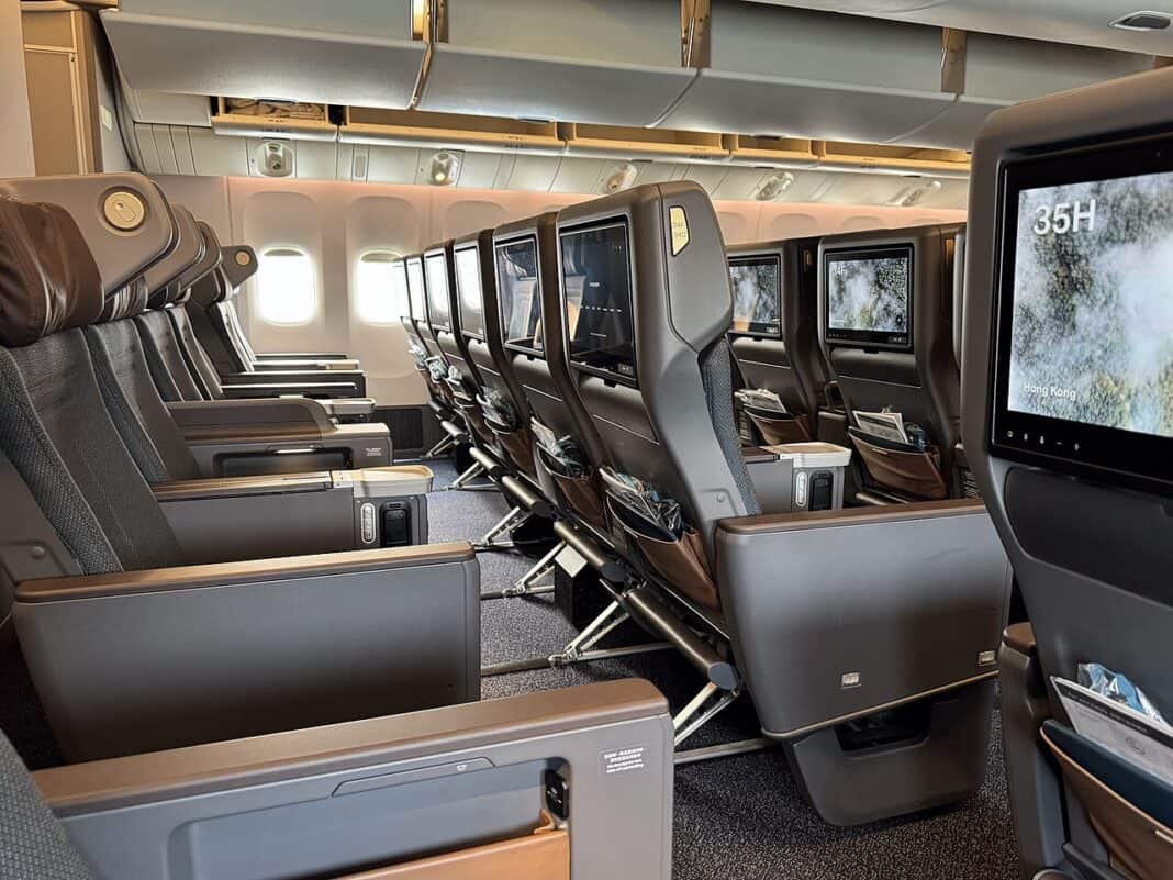 Is Premium Economy Worth It? The Honest Math, Airline by Airline Rows of brown and taupe fixed-shell premium economy recliner seats with leg rests and large seatback screens aboard a Cathay Pacific Boeing 777-300ER, one screen showing seat number 35H