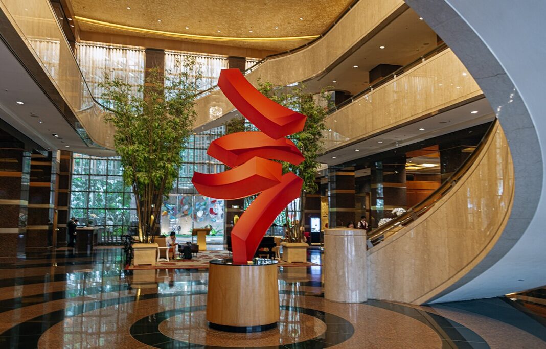 Hotel lobby with a tall red ribbon sculpture on a round plinth, polished marble floor, curving mezzanine balconies and bamboo planting behind floor-to-ceiling windows