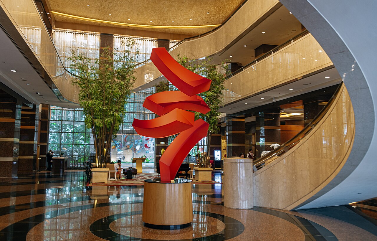 Hotel lobby with a tall red ribbon sculpture on a round plinth, polished marble floor, curving mezzanine balconies and bamboo planting behind floor-to-ceiling windows