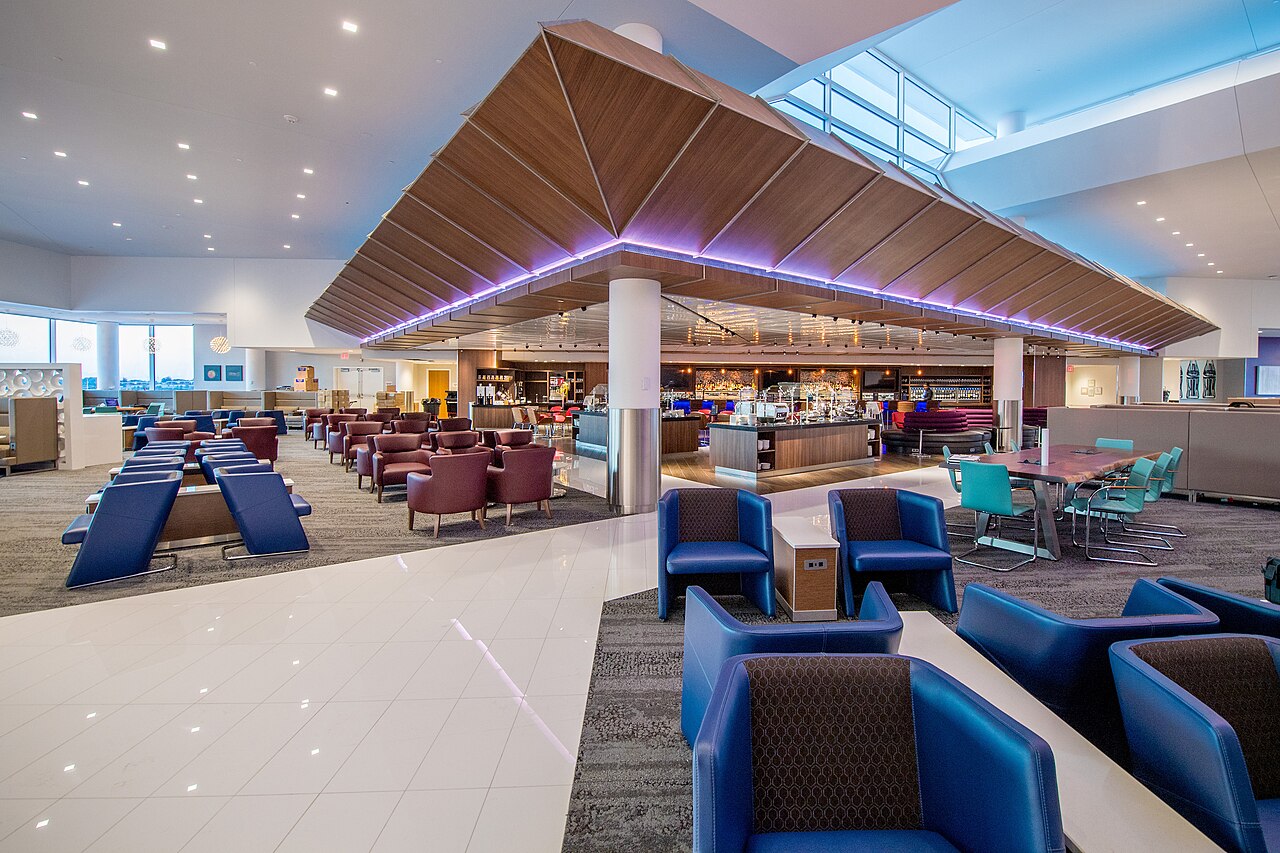 Delta Sky Club interior with blue and burgundy lounge chairs, teal meeting-table seating, a central bar and a folded wood canopy ceiling edged in purple light