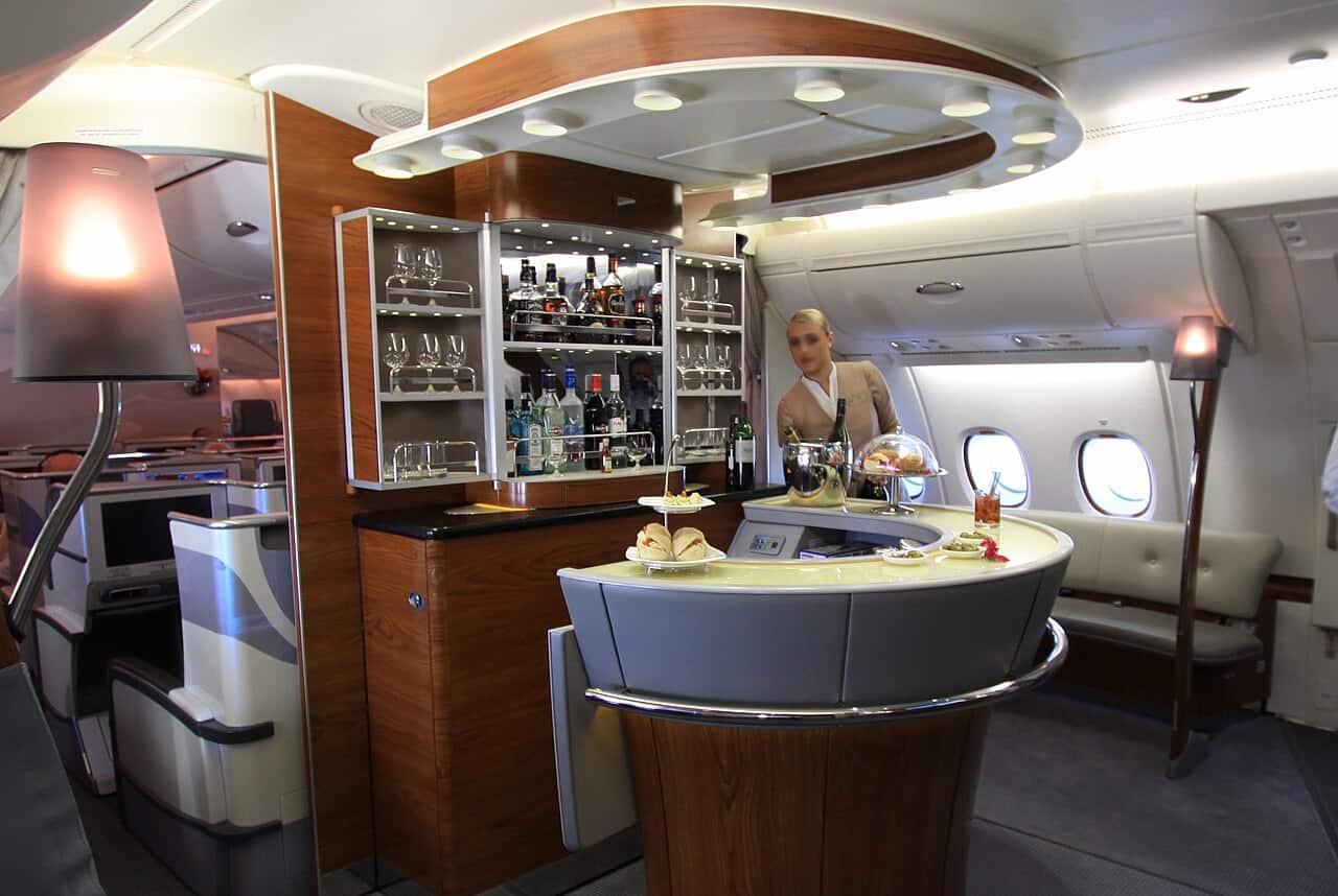 Curved onboard bar at the rear of an Emirates A380 upper deck, with a crew member serving behind it, a lit spirits cabinet, canapes and champagne on the counter and a cream leather banquette by the windows