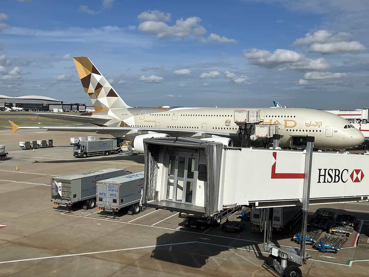 Etihad Airways Airbus A380 parked at a London Heathrow gate with a jet bridge attached, showing the airline's gold and bronze geometric tail design and Etihad titles on the fuselage