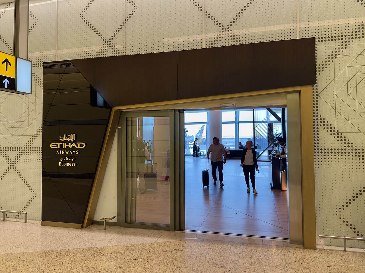 Entrance to the Etihad Airways business class lounge at Abu Dhabi airport, with gold Etihad Airways signage on a dark panel beside a glass doorway and travellers walking through the terminal beyond