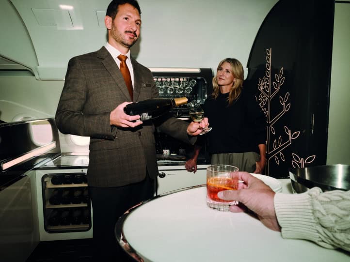 A Four Seasons crew member in a tweed suit pouring sparkling wine for a guest at the onboard bar of the Four Seasons Private Jet, beside a wine rack and a black panel with the gold Four Seasons tree motif