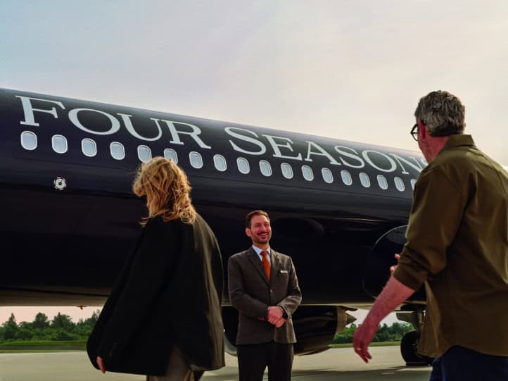 Two guests walking toward a smiling Four Seasons staff member in a tweed suit on the tarmac, beside the black fuselage of the Four Seasons Private Jet with large silver Four Seasons lettering