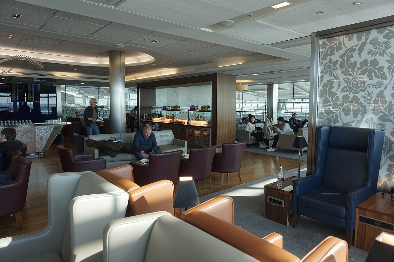 Airport club lounge interior with leather armchairs in tan, burgundy and navy, a self-serve spirits bar, coffee station and departure boards visible against the terminal windows