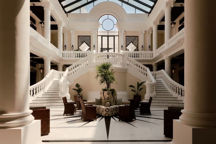 White neoclassical hotel lobby atrium with a sweeping double staircase, columns, glass skylight ceiling and rattan armchairs around a patterned planter