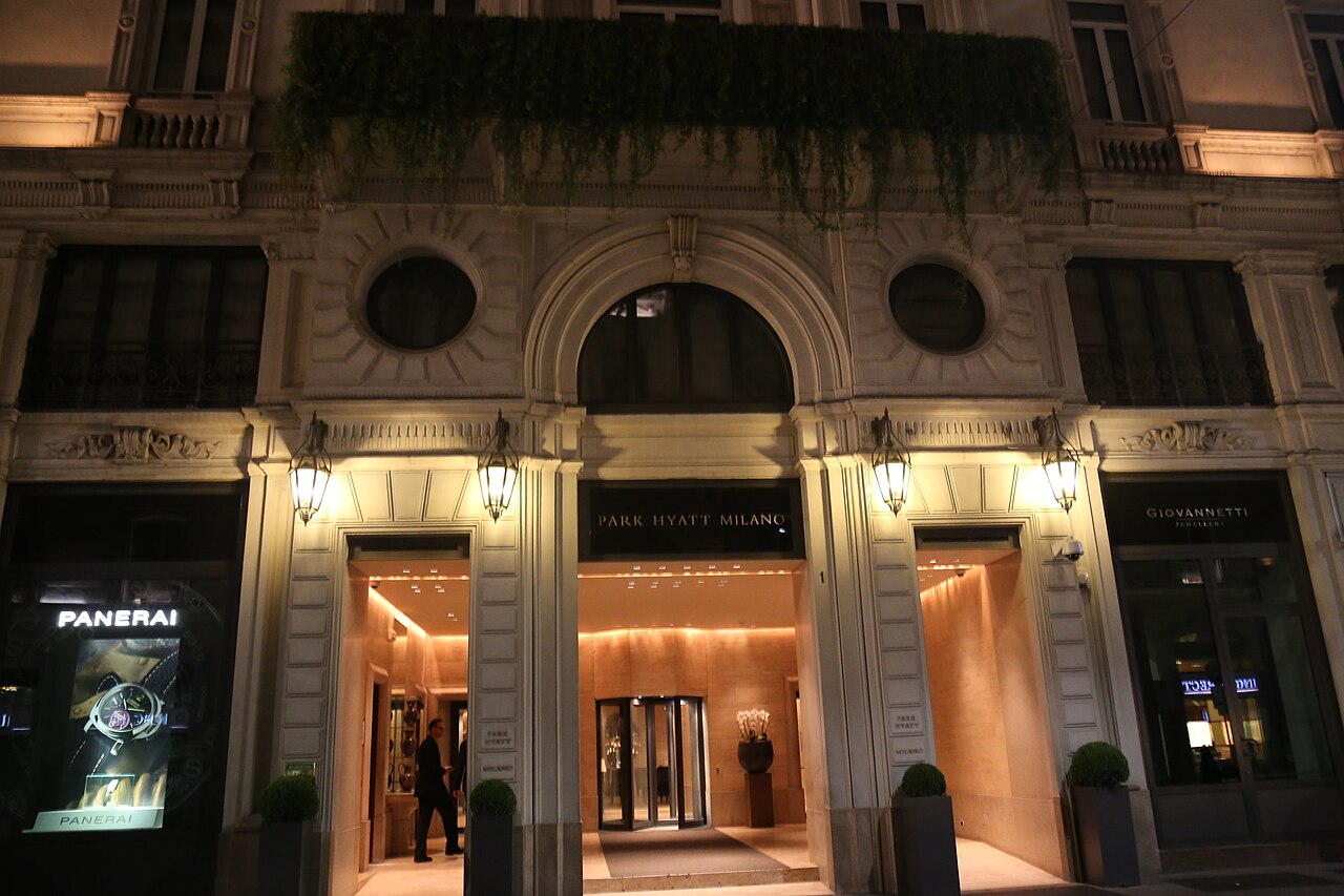 Stone arched entrance of the Park Hyatt Milano lit at night, with PARK HYATT MILANO lettering above a revolving door, hanging lanterns, trailing greenery on the balcony and a luxury boutique window to one side
