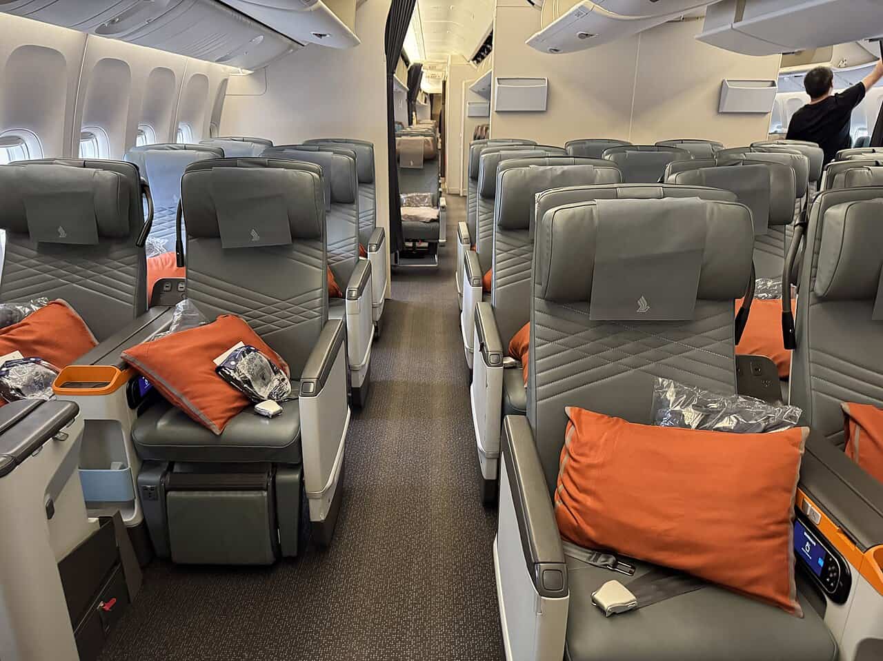 Singapore Airlines premium economy cabin with grey leather recliner seats, orange pillows and amenity packs on the seats, arranged two-four-two with the airline's bird logo on the headrest covers