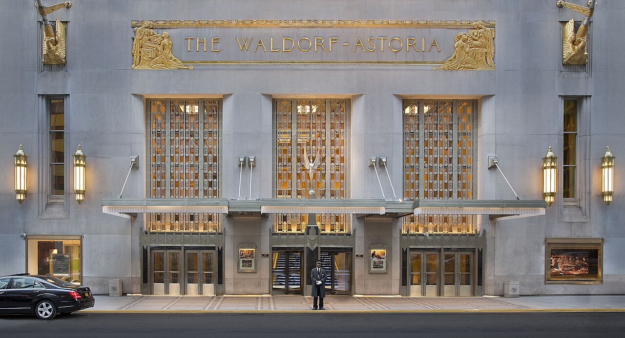 Art Deco limestone facade of the Waldorf Astoria New York with gilded relief panels, gold lettering reading THE WALDORF-ASTORIA, lit lanterns and a uniformed doorman under the Park Avenue canopy