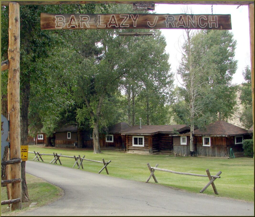 All-Inclusive Resorts in the USA: What Actually Exists in 2026 A timber archway carved with the words Bar Lazy J Ranch over a gravel drive, with log cabins, a split-rail fence and tall cottonwoods behind it