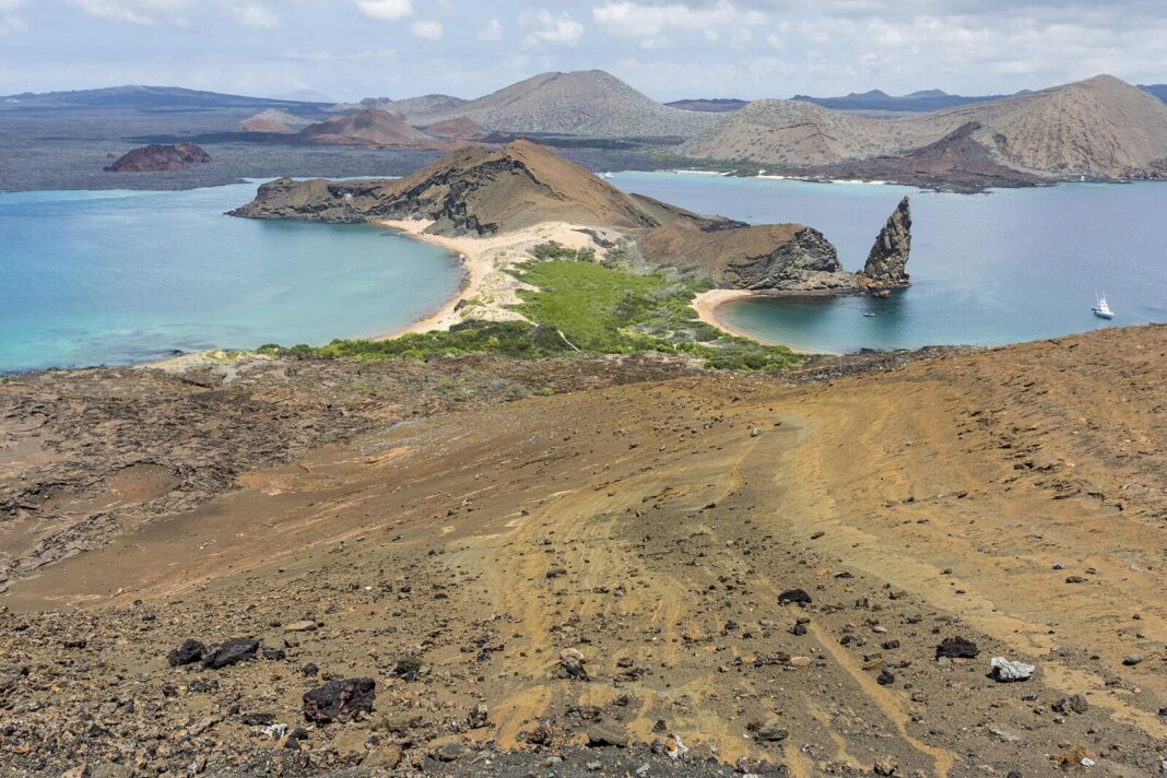 Aerial view of Bartolome Island showing the jagged spire of Pinnacle Rock, two crescent beaches meeting at a narrow green isthmus, turquoise bays on either side, volcanic cones in the distance and a white yacht anchored offshore