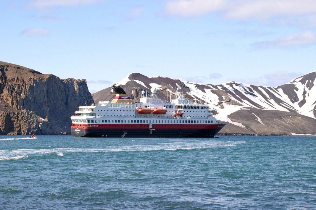 Luxury Antarctica Cruise 2026-27: Ships, Real Prices and How to Choose A white expedition cruise ship with a red hull stripe and orange lifeboats anchored in calm green-blue water below steep brown volcanic cliffs streaked with snow, with a small red inflatable boat running alongside