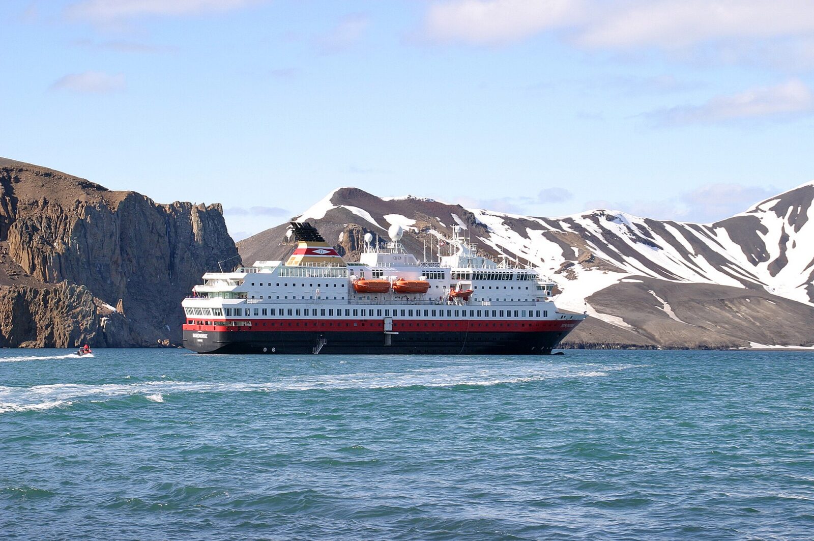 A white expedition cruise ship with a red hull stripe and orange lifeboats anchored in calm green-blue water below steep brown volcanic cliffs streaked with snow, with a small red inflatable boat running alongside