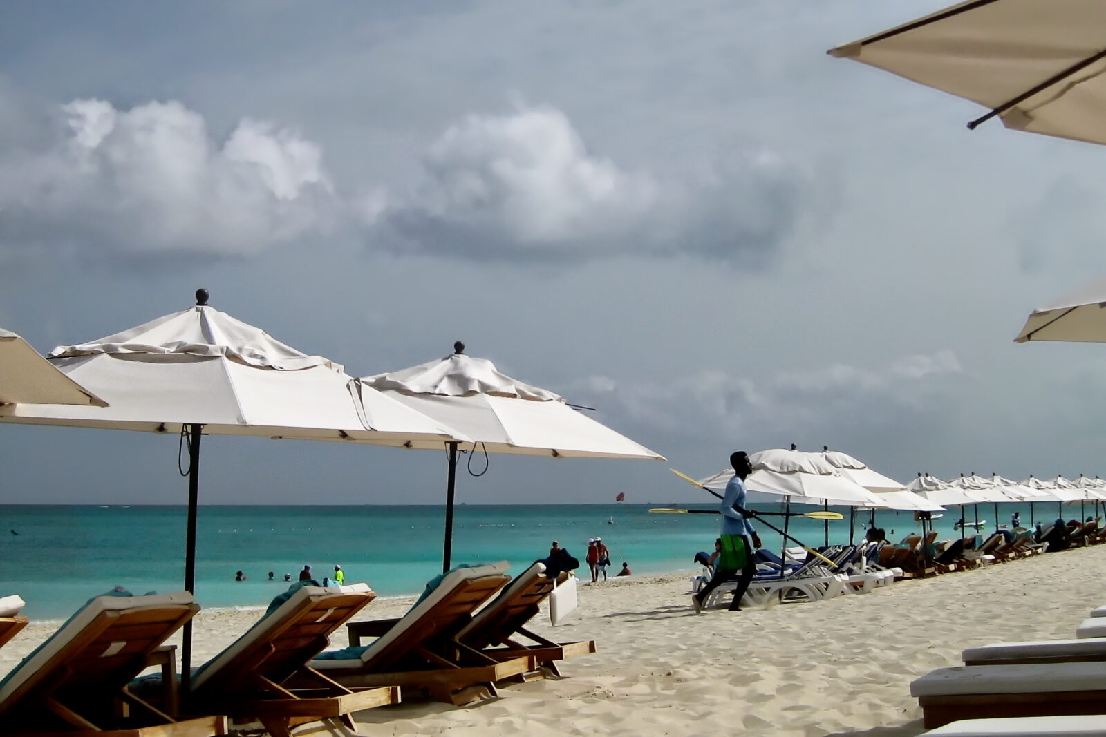 A long row of white canvas umbrellas and wooden sun loungers on the white sand at Grace Bay, Turks and Caicos, with swimmers in turquoise water and a beach attendant carrying a yellow-bladed paddle