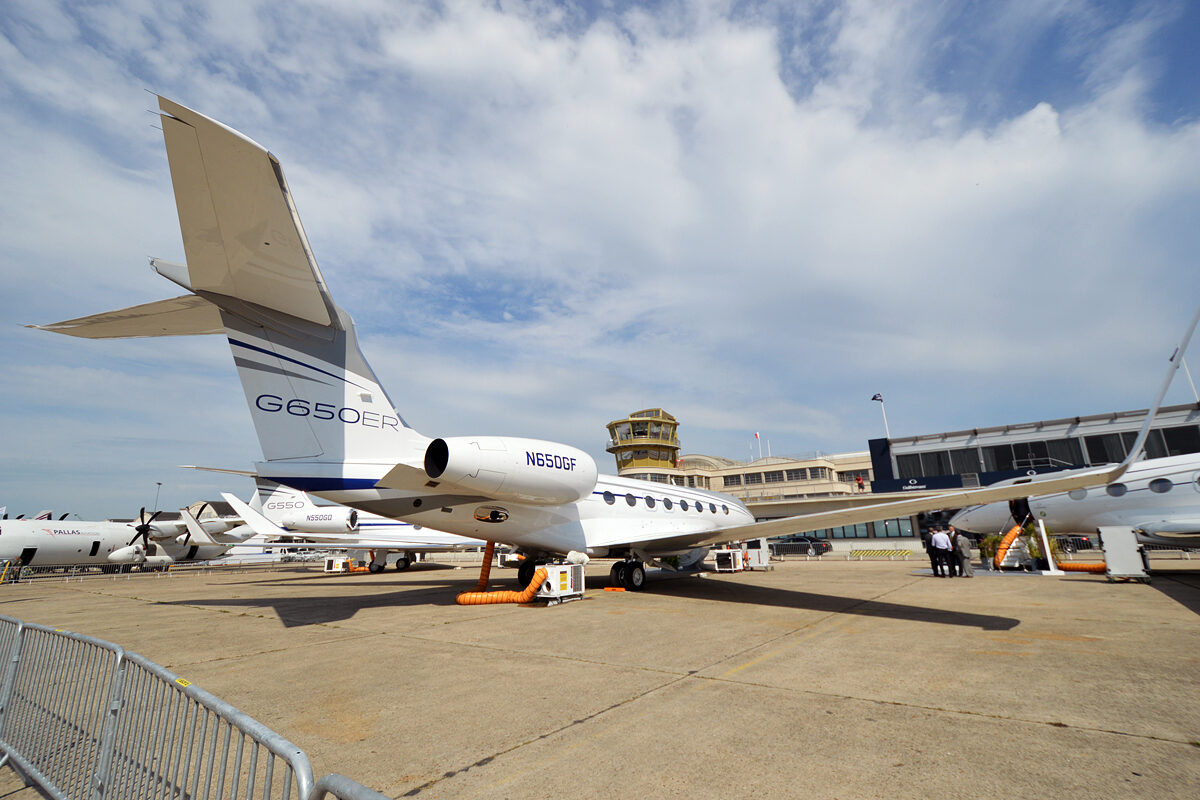 A white Gulfstream G650ER registered N650GF parked on an airport apron beside a Gulfstream G550, both with orange ground-conditioning hoses running to the fuselage, with a control tower and terminal building behind them