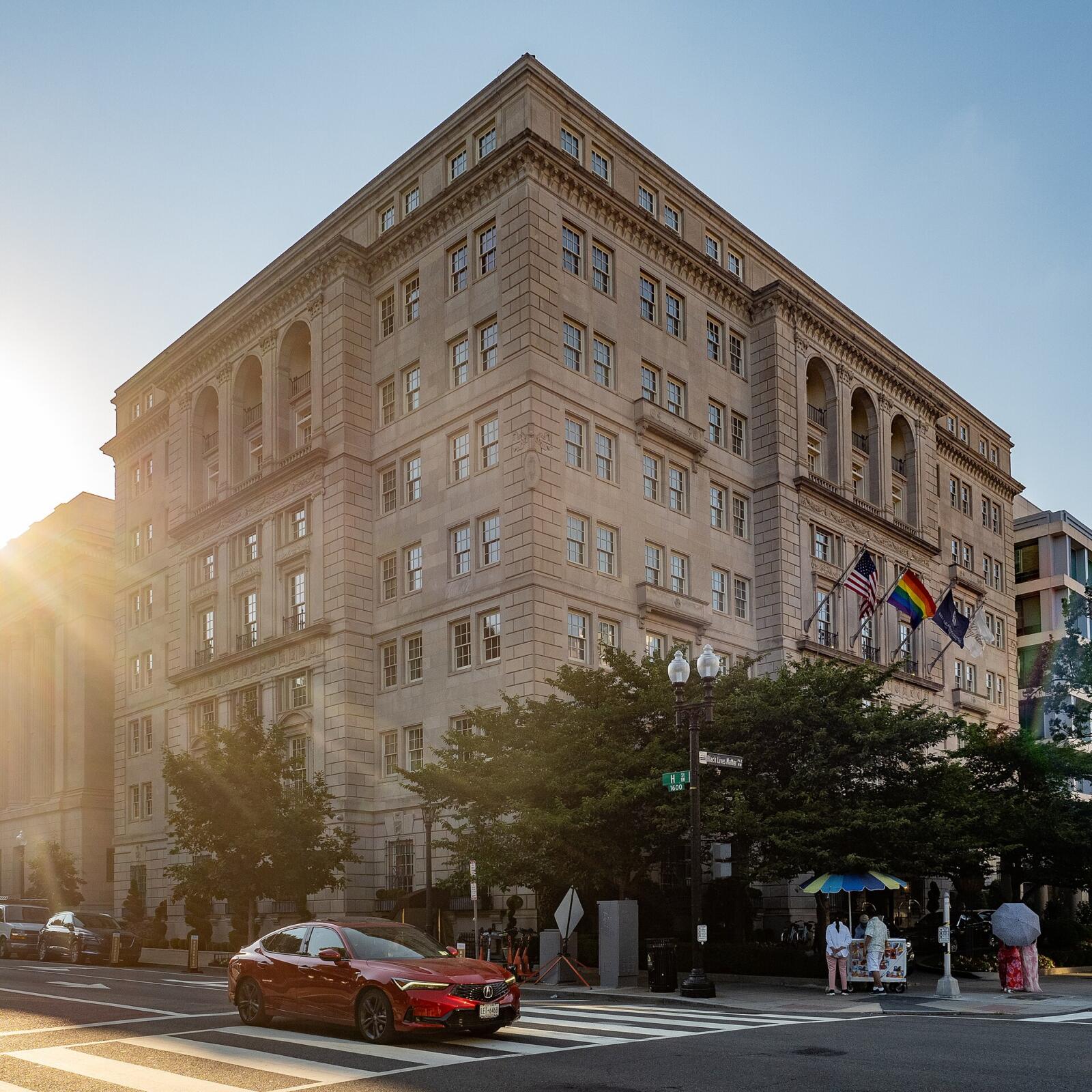 The limestone corner facade of the Hay-Adams hotel in Washington DC at low sun, seven storeys with a carved cornice and arched loggia windows at the top floor, US, rainbow and hotel flags above the entrance, and street signs reading H St 1600 and Black Lives Matter Plaza at the crosswalk below.