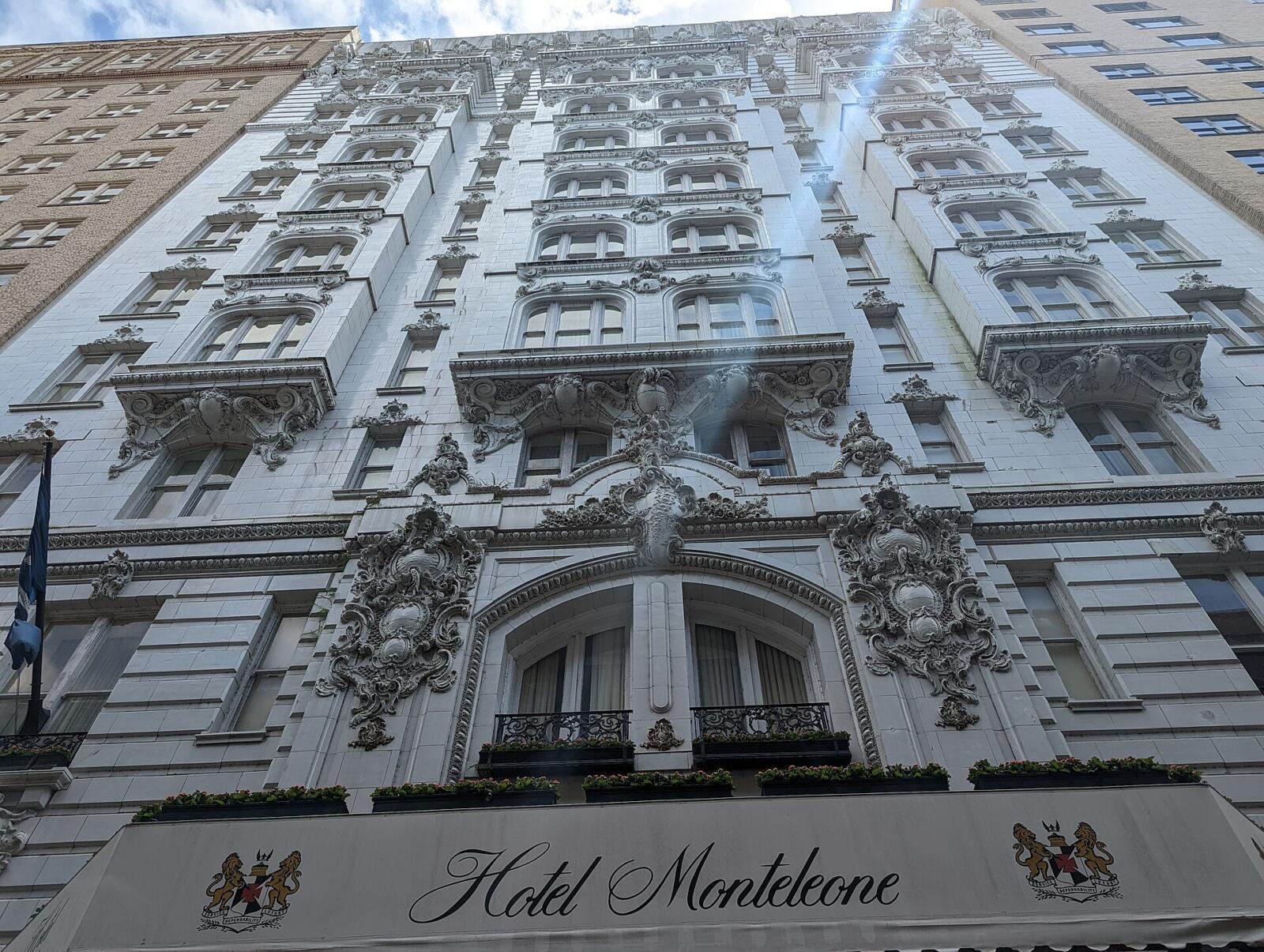 The white Beaux-Arts facade of the Hotel Monteleone on Royal Street in New Orleans, photographed steeply upward: ornate carved cartouches and scrollwork around the windows, wrought-iron balconies with pink and white flower boxes, and a white entrance awning lettered Hotel Monteleone in script between two gold lion-and-shield crests.