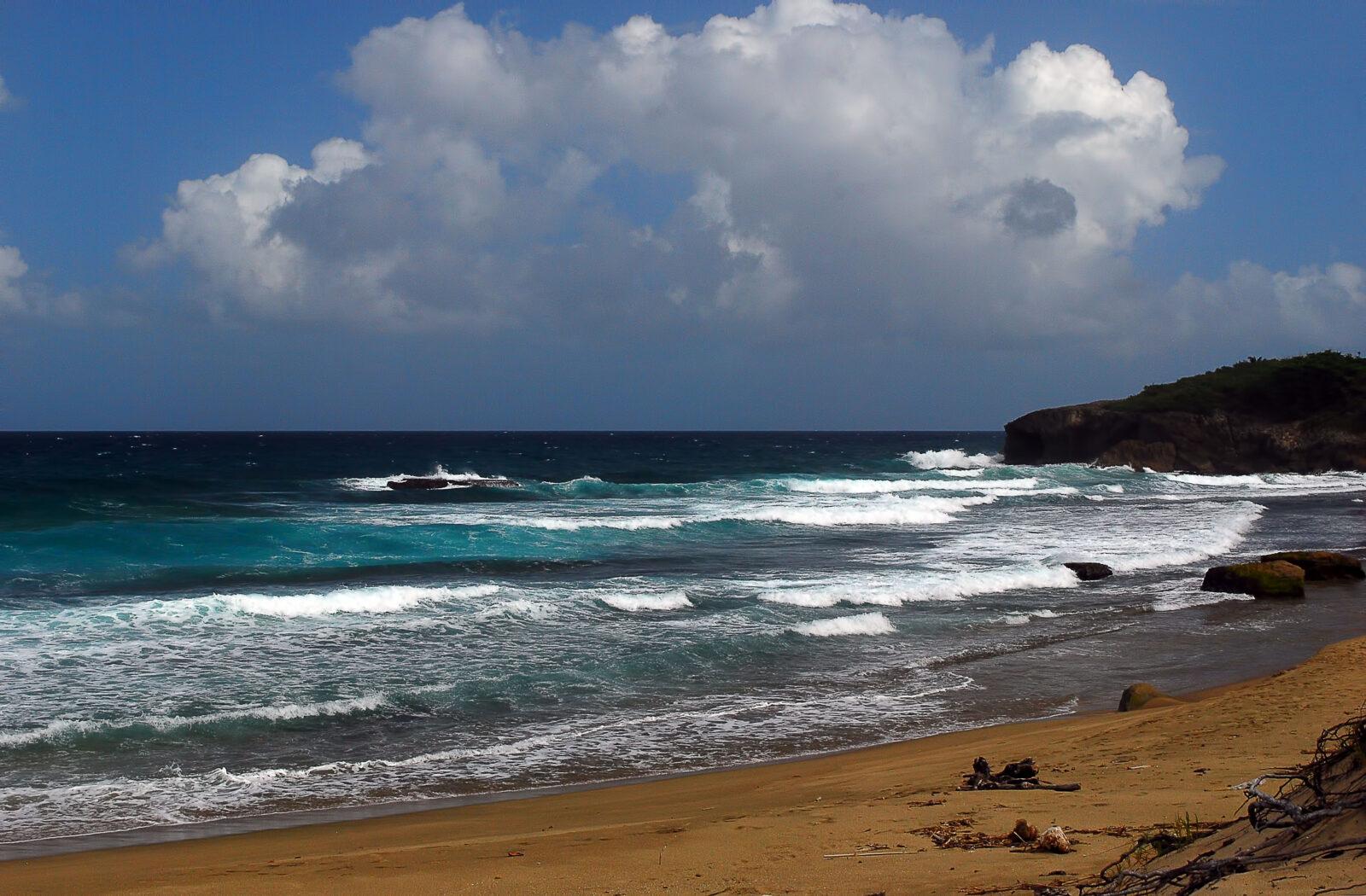 Rows of whitecaps breaking onto a tan sand beach at Jobos Beach in Isabela, Puerto Rico, with a green-topped limestone headland at right and tall cumulus clouds overhead