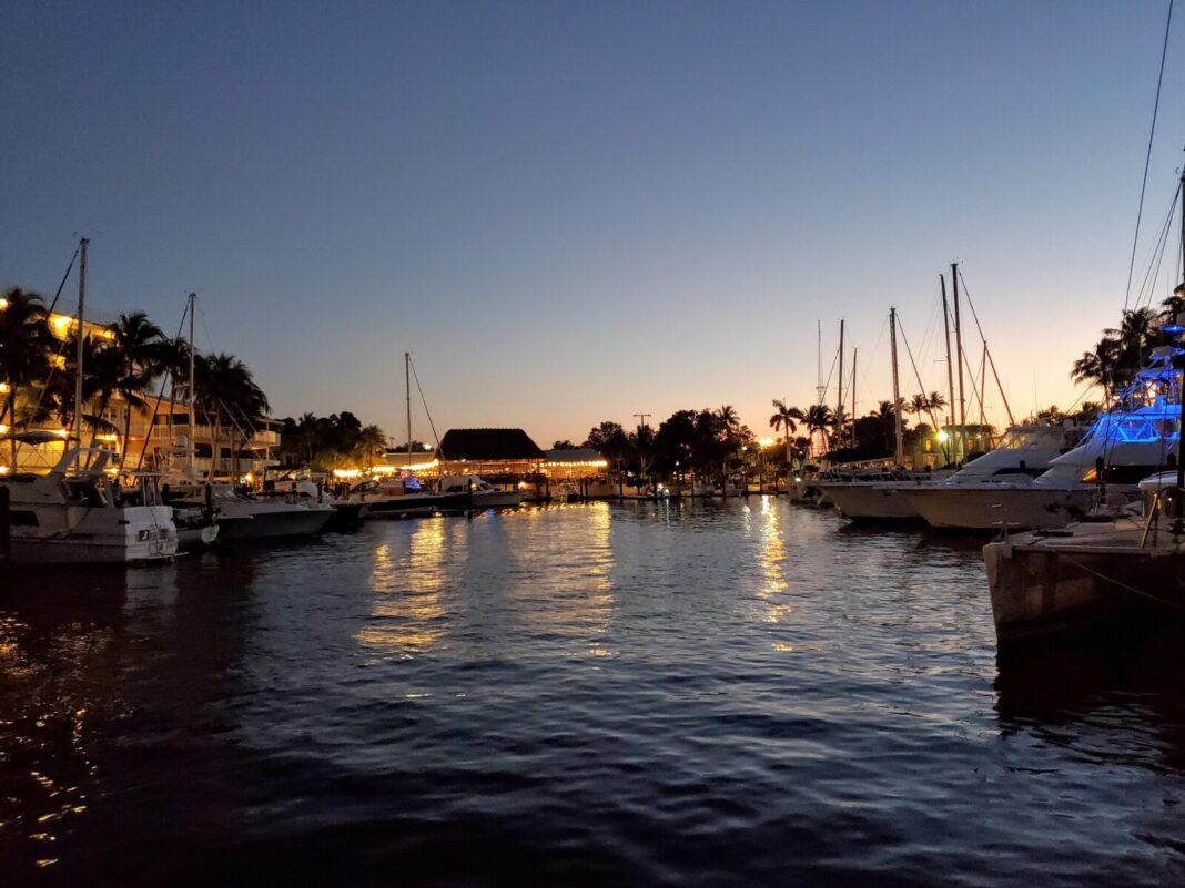 Sportfishing boats and sailboats moored either side of a channel at a Key Largo marina at dusk, with a lit tiki-roofed waterfront restaurant, palms and a balconied resort building reflected in the water