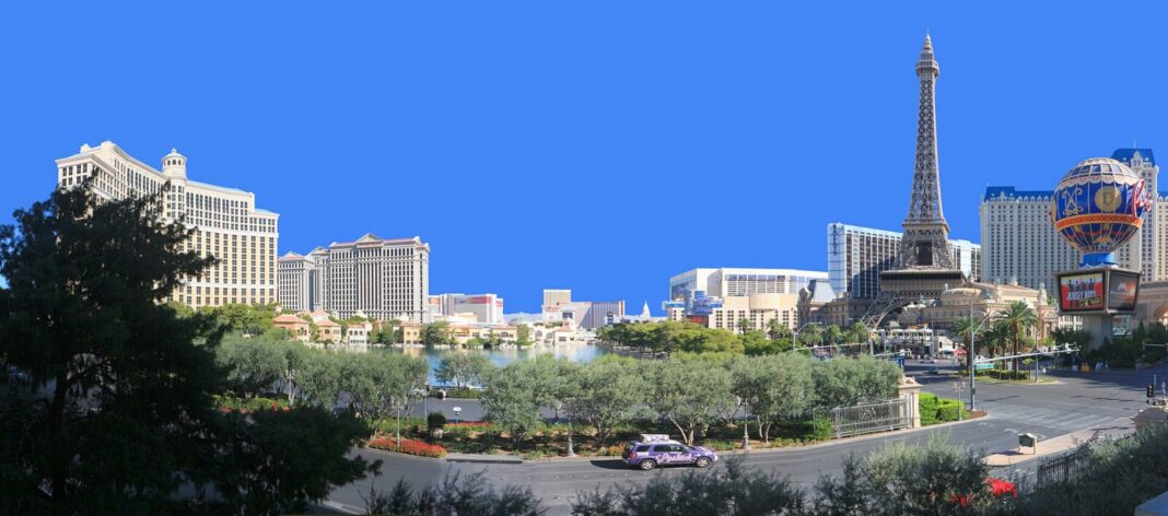 Luxury Hotels in Las Vegas 2027: Only 3 Hold Five Diamond — Here’s the Real List Daytime panorama of the Las Vegas Strip under a deep blue sky: the cream towers of Bellagio and its lake on the left, the Caesars Palace towers in the centre, and Paris Las Vegas with its half-scale Eiffel Tower and hot-air-balloon sign on the right, with olive trees along the boulevard in the foreground.